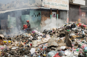 En una calle de Puerto Príncipe, una pila de basura incendiándose, un hombre en motocicleta, y un edificio en ruinas al fondo.