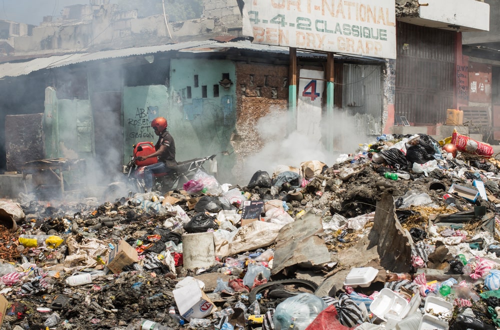 En una calle de Puerto Príncipe, una pila de basura incendiándose, un hombre en motocicleta, y un edificio en ruinas al fondo.