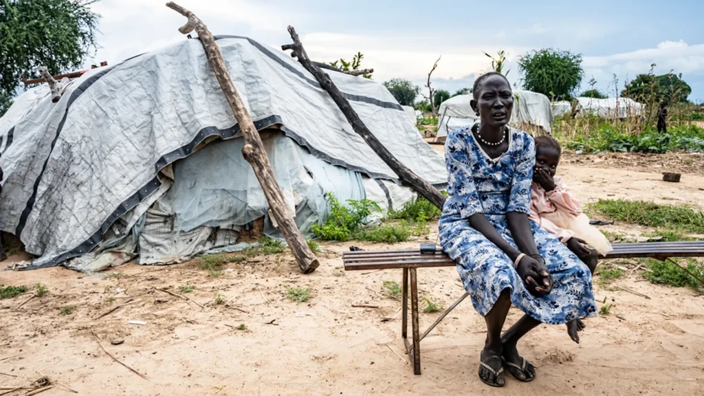 Arual Manyok y su hija Adit Ayuel se sientan en un banco frente a su refugio en un campamento de desplazados. Sudán del Sur, octubre de 2024.