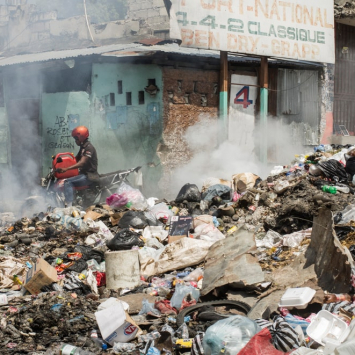En una calle de Puerto Príncipe, una pila de basura incendiándose, un hombre en motocicleta, y un edificio en ruinas al fondo.