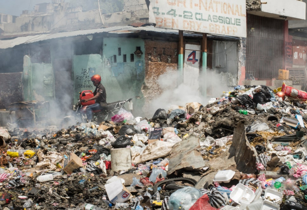 En una calle de Puerto Príncipe, una pila de basura incendiándose, un hombre en motocicleta, y un edificio en ruinas al fondo.