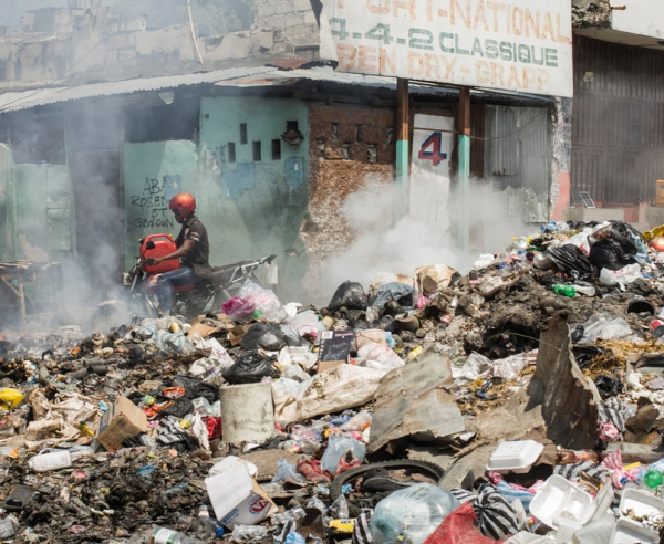En una calle de Puerto Príncipe, una pila de basura incendiándose, un hombre en motocicleta, y un edificio en ruinas al fondo.