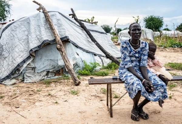 Arual Manyok y su hija Adit Ayuel se sientan en un banco frente a su refugio en un campamento de desplazados. Sudán del Sur, octubre de 2024.