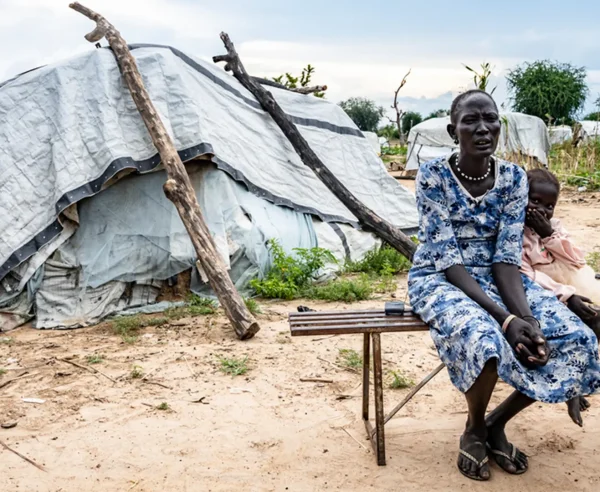 Arual Manyok y su hija Adit Ayuel se sientan en un banco frente a su refugio en un campamento de desplazados. Sudán del Sur, octubre de 2024.