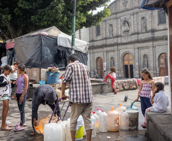 Personas migrantes atrapados entre la violencia, las estrategias de disuasión y los asfixiantes procedimientos de asilo. Un grupo de niños y adultos recoge agua en la Plaza de la Soledad, en la Ciudad de México.